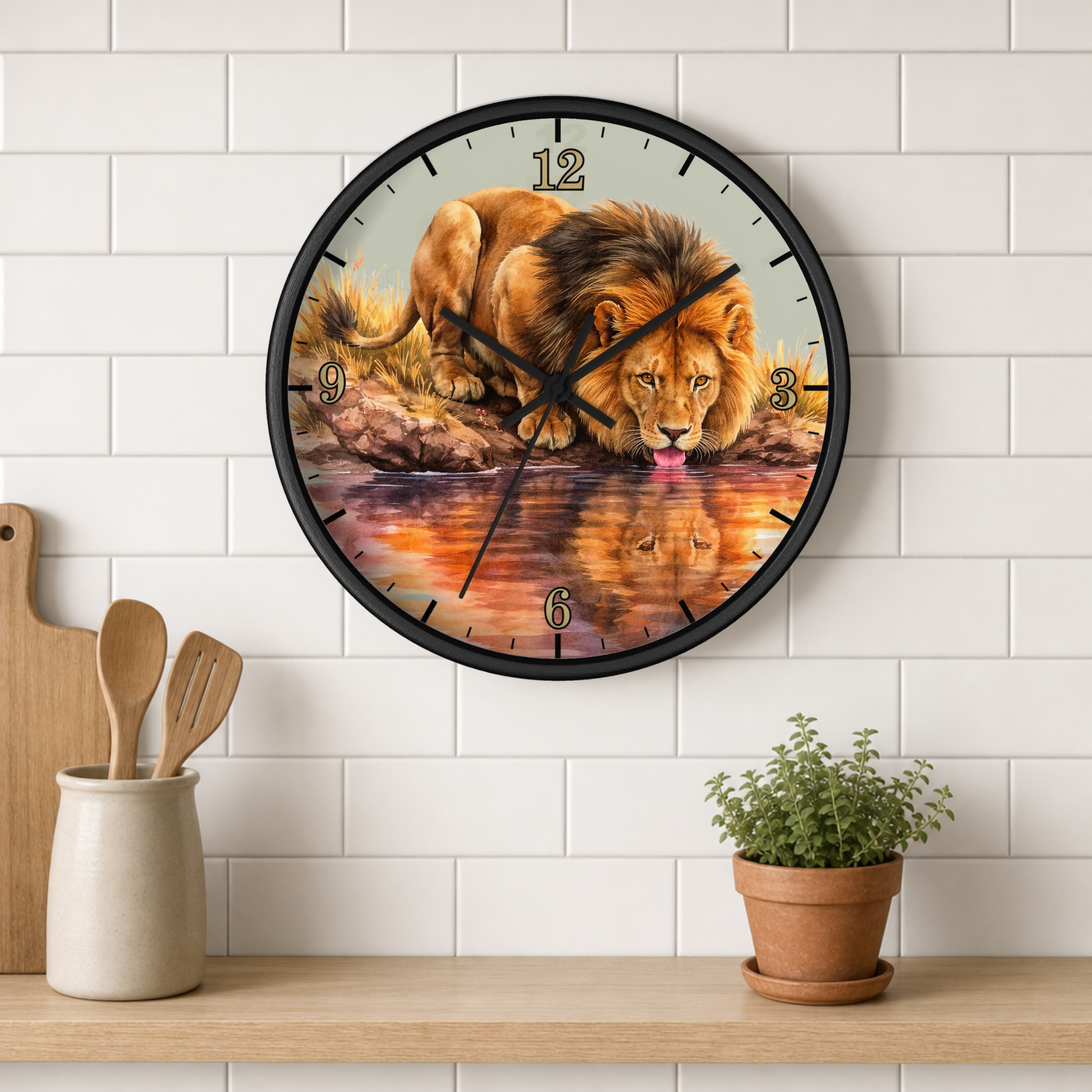 A round wall clock with a black frame and a printed lion drinking water design, mounted on a white subway tile wall above a wooden shelf with kitchen utensils and a potted plant.