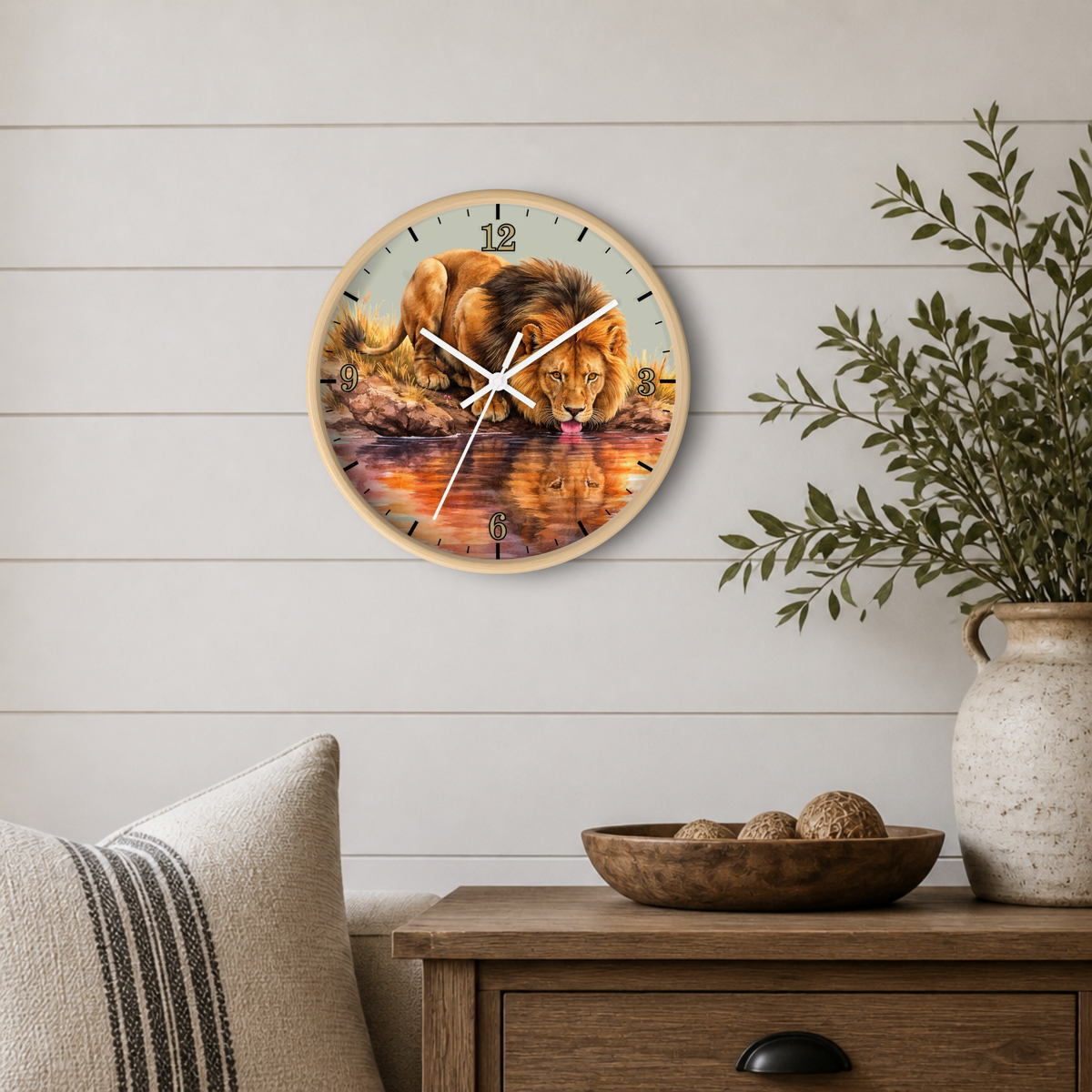 A round wall clock with a natural wood frame hangs on a white shiplap wall above a wooden side table. The clock face features a detailed painting of a lion drinking water from a reflective pool, with black Arabic numerals and white clock hands.