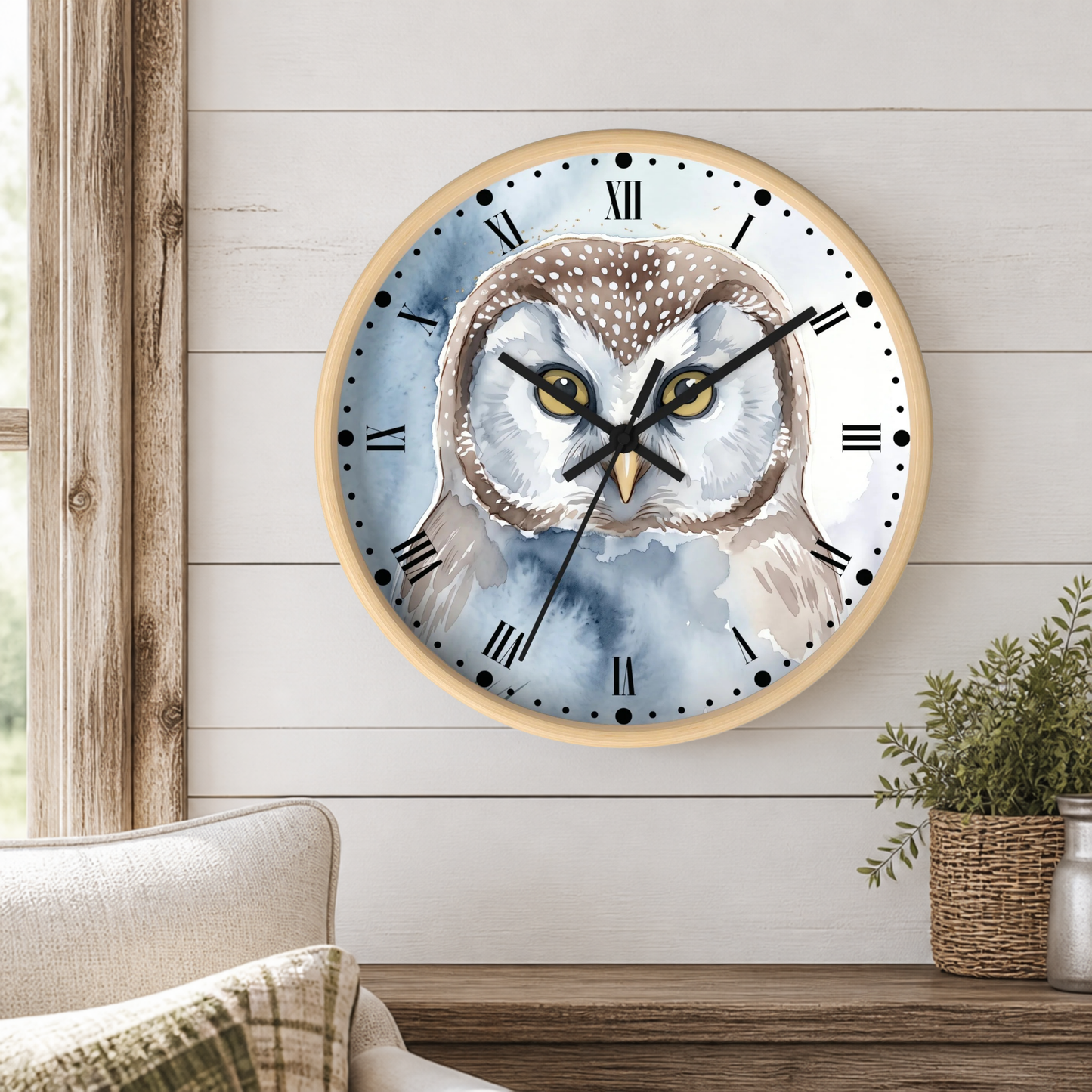 A round wall clock with a watercolor owl illustration on the face, mounted on a white shiplap wall above a wooden table with a potted plant and part of a sofa visible.