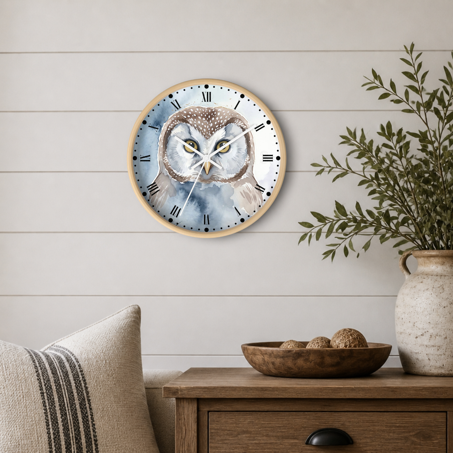 Round wall clock with a watercolor owl design on the face, mounted on a white shiplap wall above a wooden side table with a vase and bowl.