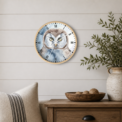 Round wall clock with a watercolor owl design on the face, mounted on a white shiplap wall above a wooden side table with a vase and bowl.