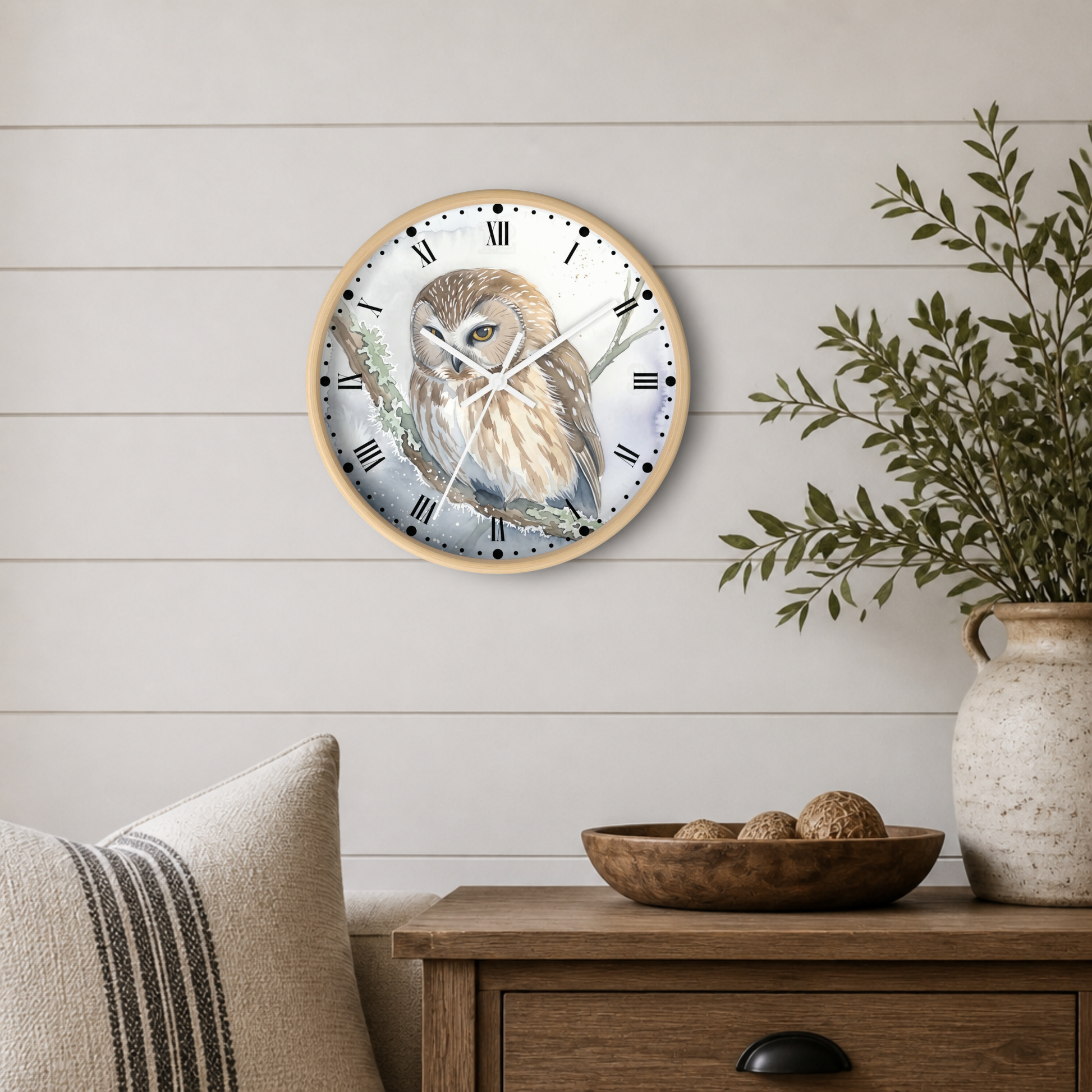 A round wall clock with a light wood frame hangs on a white shiplap wall above a wooden side table. The clock face features a detailed illustration of a brown and cream owl perched on a branch.