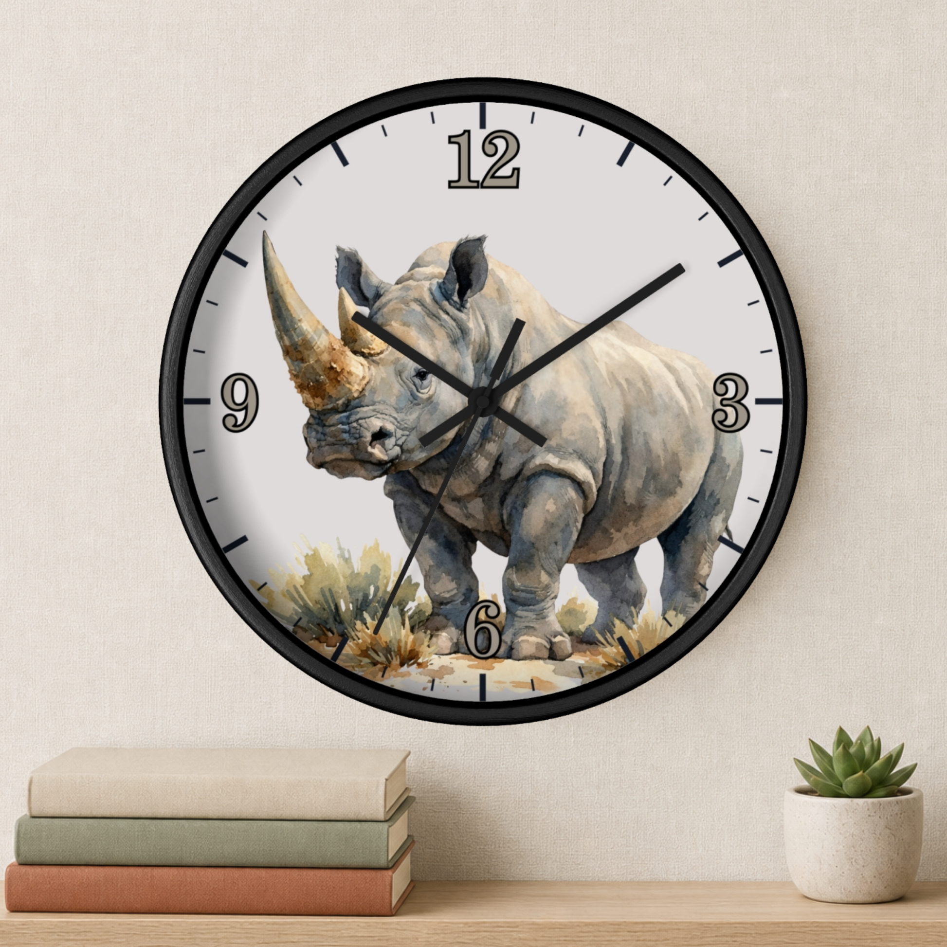 A round wall clock with a black frame and a watercolor-style rhinoceros illustration on the face, mounted on a light beige wall above a shelf with stacked books and a small potted succulent.