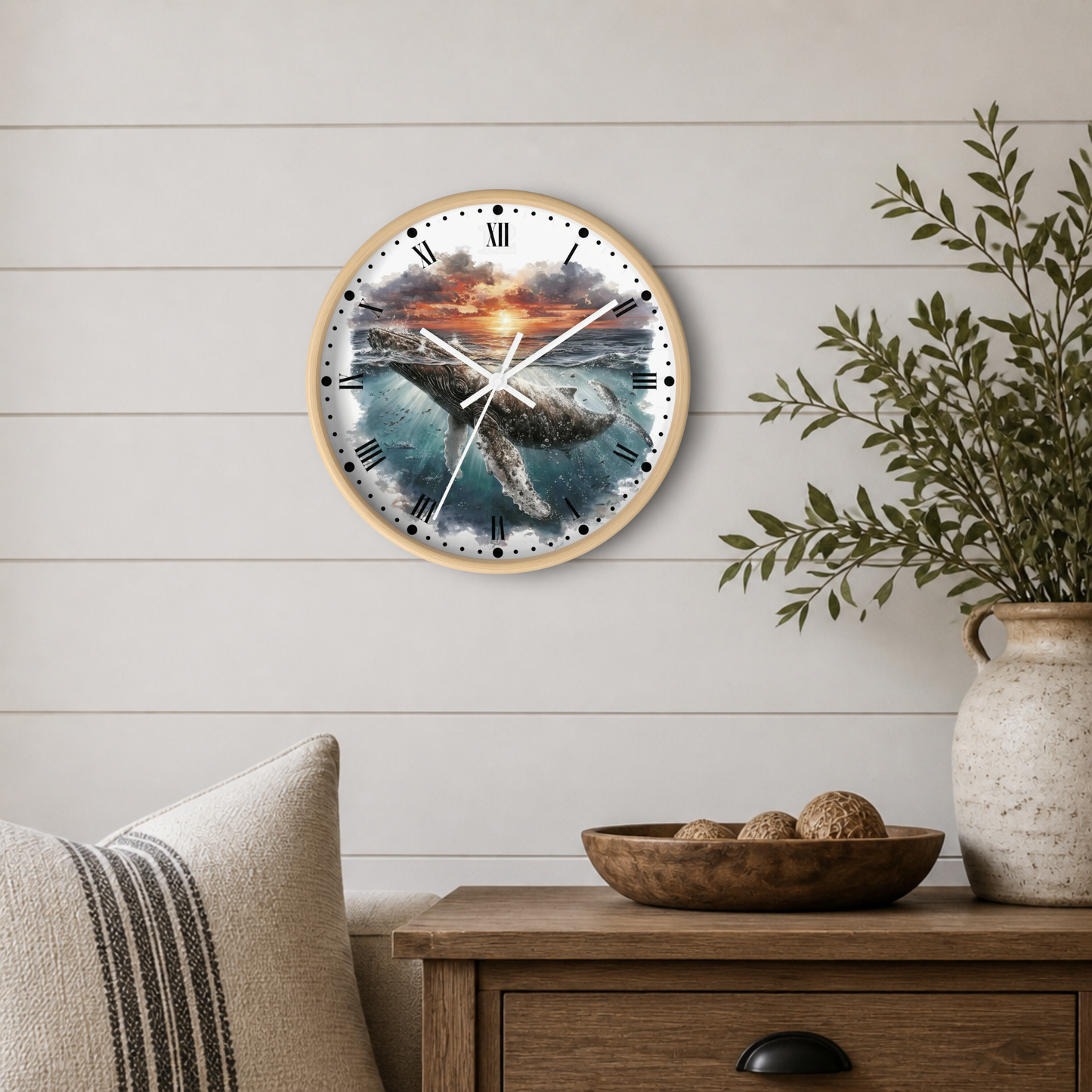 A round wall clock with a wood frame hangs on a white shiplap wall above a wooden side table. The clock face features a dramatic whale swimming through blue ocean water at sunset, with black Roman numerals and white clock hands.