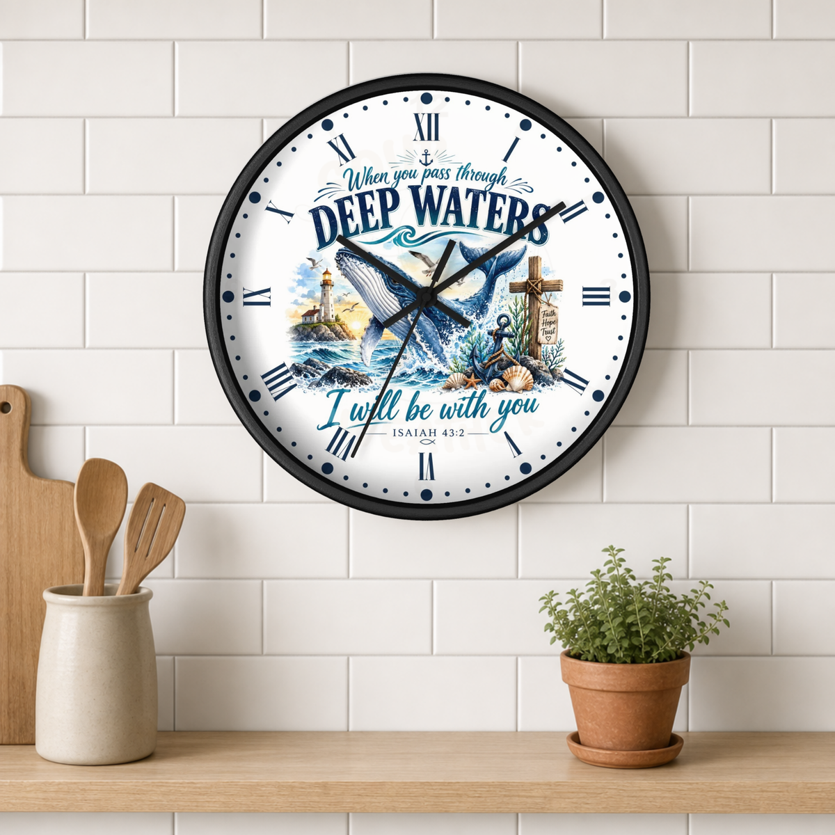 A round wall clock with a black frame hangs on a white subway tile wall above a wooden shelf. The clock face features a blue whale, a lighthouse, a cross, and the text 'When you pass through DEEP WATERS I will be with you ISAIAH 43:2'.