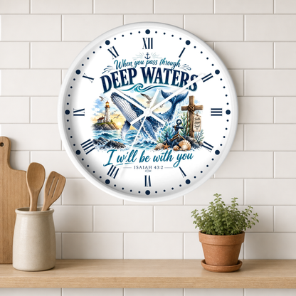 A round wall clock with a white face and Roman numerals, featuring a nautical illustration of a whale, lighthouse, anchor, and cross, with the text 'When you pass through DEEP WATERS I will be with you ISAIAH 43:2'. The clock is shown hanging on a white subway tile wall above a wooden shelf with a small potted plant and wooden utensils.