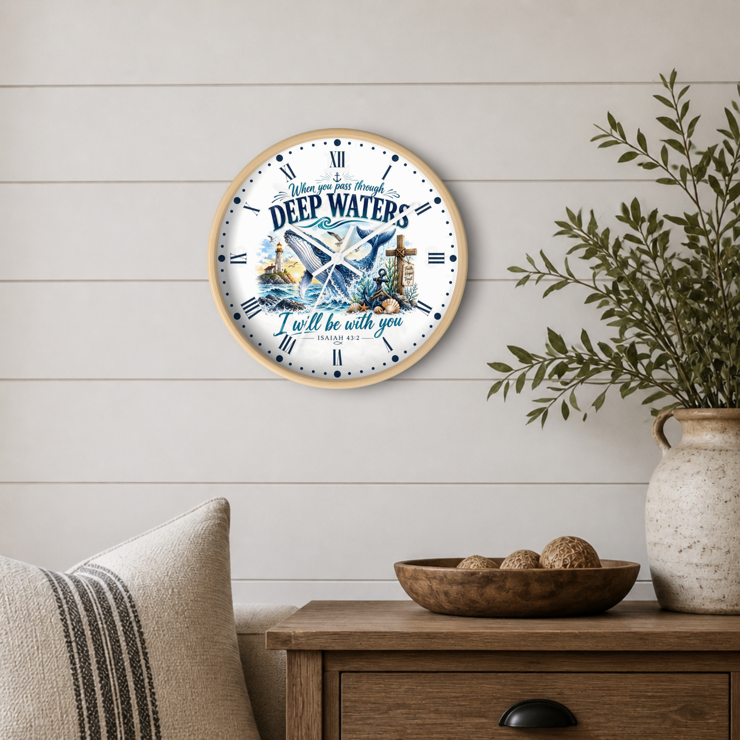 A round wall clock with a light wood frame hangs on a white shiplap wall above a wooden side table. The clock face features a nautical illustration of a blue whale, a lighthouse, and the text 'When you go through DEEP WATERS I'll be with you'.