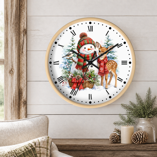 A round wall clock with a light wood frame hangs on a white shiplap wall above a wooden table. The clock face features a festive Christmas illustration with a snowman in a red knit hat and scarf, a deer, evergreen trees, and a basket of red bows.