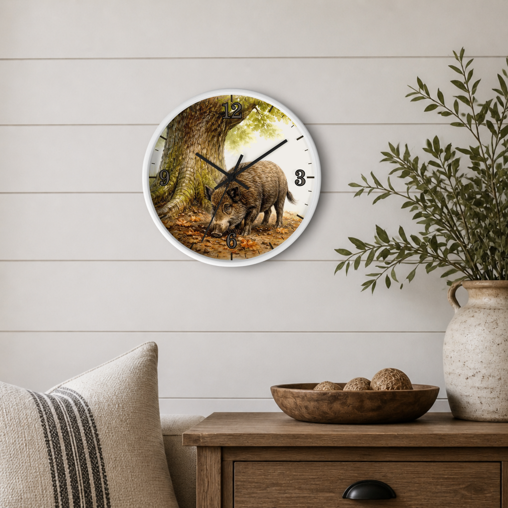 Round wall clock with a white frame featuring a detailed illustration of a wild boar in front of a tree, styled on a light wood paneled wall above a wooden side table with a plant and decorative bowl.