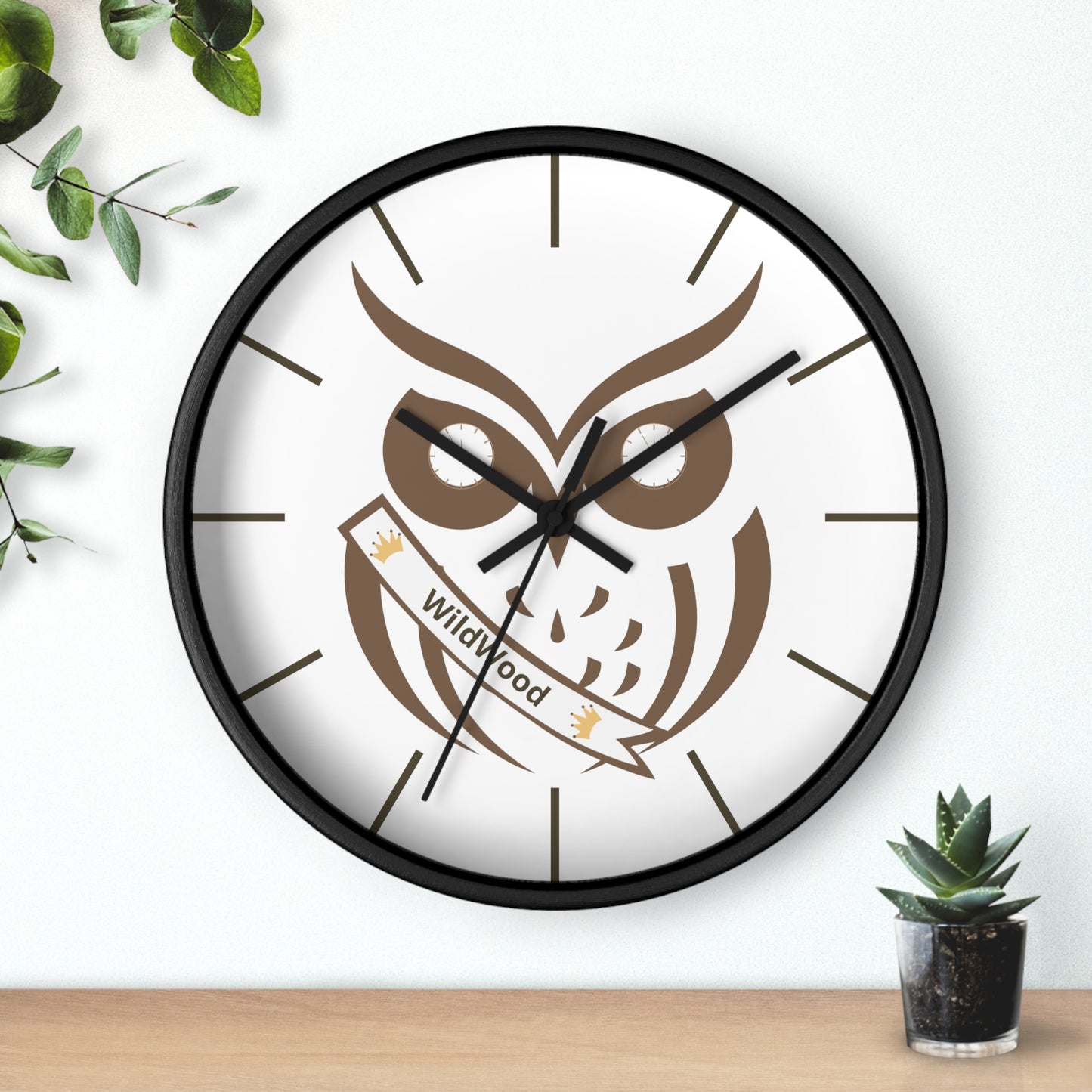 A round wall clock with a black frame and a white face featuring a brown owl graphic and a banner that reads 'Wildwood'. The clock is mounted on a white wall above a light wood surface, with green leaves and a small potted succulent as decorative props.