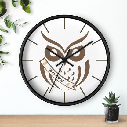 A round wall clock with a black frame and a white face featuring a brown owl graphic and a banner that reads 'Wildwood'. The clock is mounted on a white wall above a light wood surface, with green leaves and a small potted succulent as decorative props.