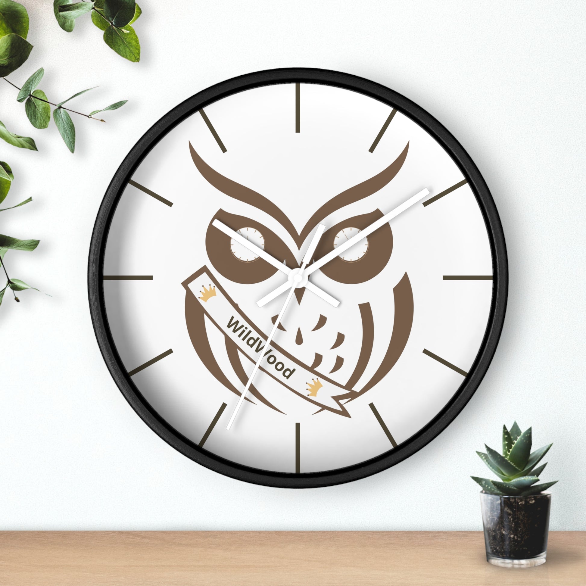 A round wall clock with a black frame and a white face featuring a brown owl illustration and a banner that reads 'Wild Good'. The clock is mounted on a white wall above a light wood surface, with green leaves and a small potted succulent as decorative props.