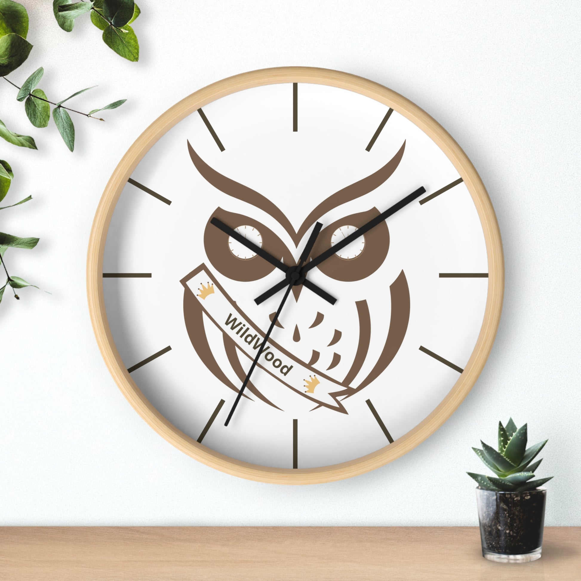A round wall clock with a light wood frame and a white face featuring a large brown owl graphic and the words 'Wildwood' on a banner. The clock is mounted on a white wall above a light wood surface, with decorative green leaves on the left and a small potted succulent on the right.