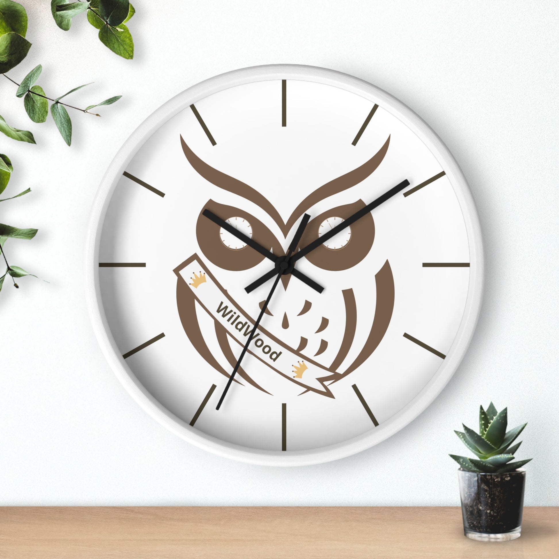 A round wall clock with a white face and white frame, featuring a brown owl graphic holding a banner that reads 'Wildwood'. The clock is styled on a light wall above a wooden surface, with green leaves and a small succulent plant as props.