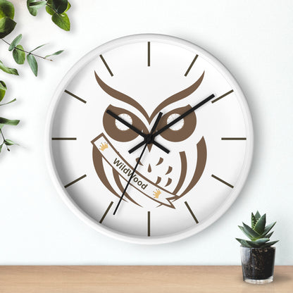 A round wall clock with a white face and white frame, featuring a brown owl graphic holding a banner that reads 'Wildwood'. The clock is styled on a light wall above a wooden surface, with green leaves and a small succulent plant as props.