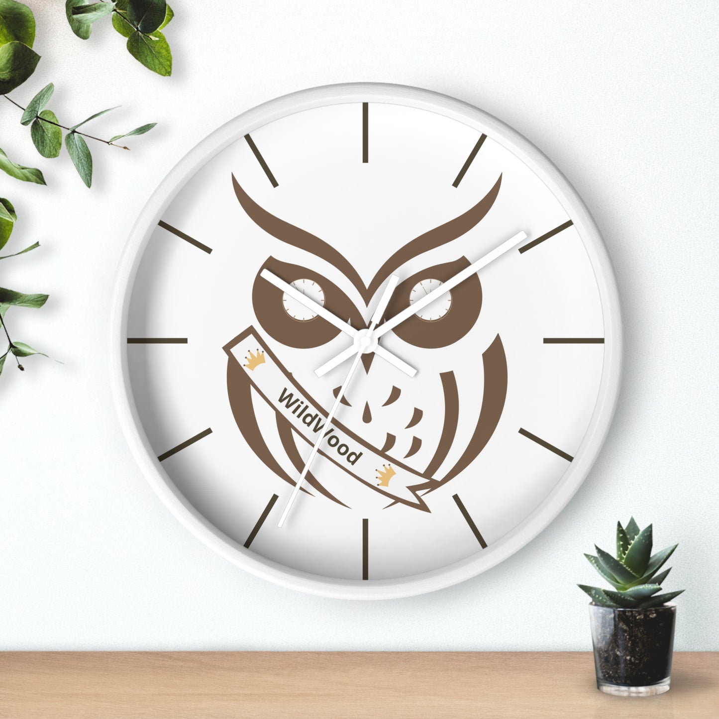 A round wall clock with a white face and white frame, featuring a brown owl illustration holding a banner that reads 'Wild Food'. The clock is mounted on a white wall above a light wood surface, with green leaves and a small succulent plant as decorative props.