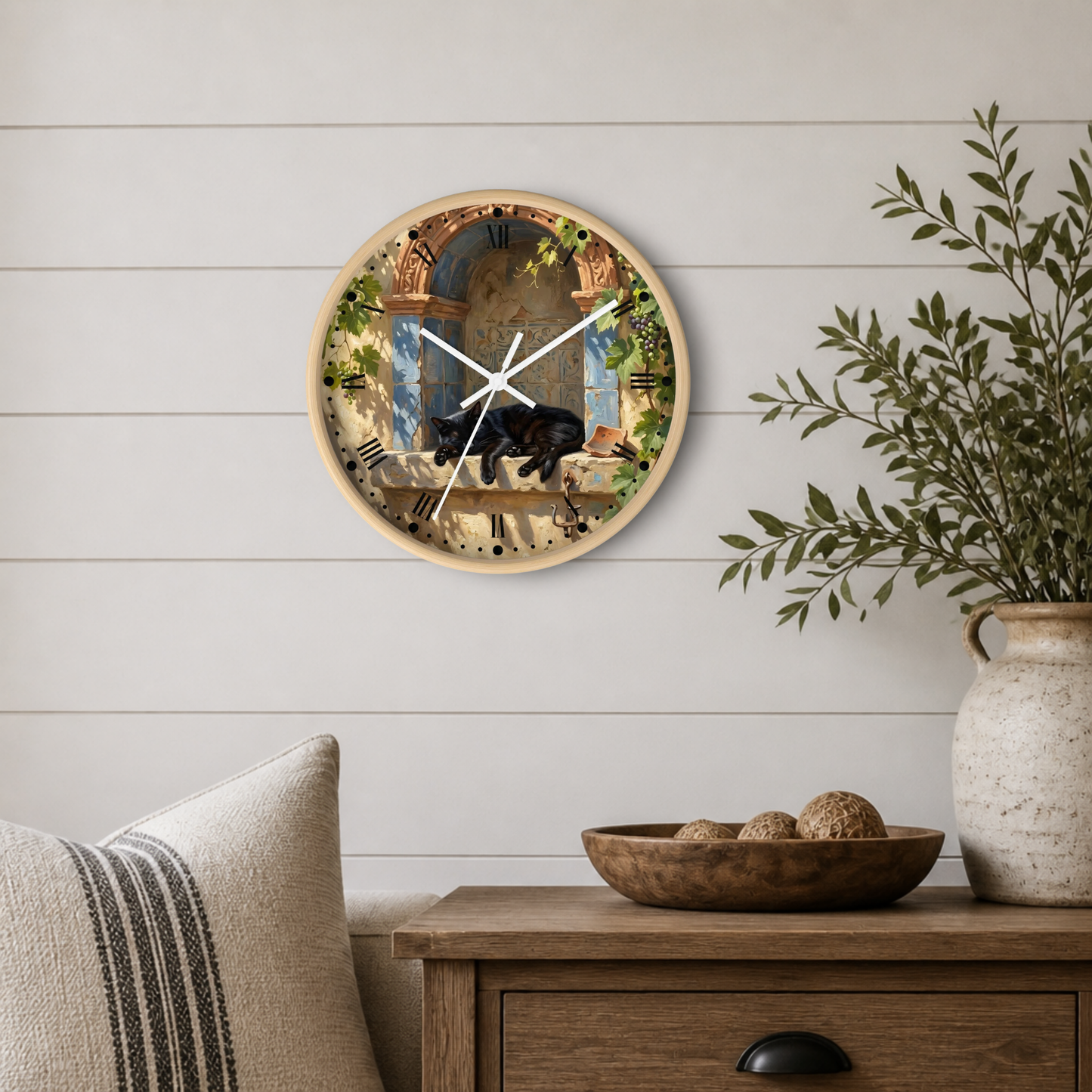A round wall clock with a light wood frame hangs on a white shiplap wall above a wooden side table. The clock face features a classical painting of a black cat in a garden.