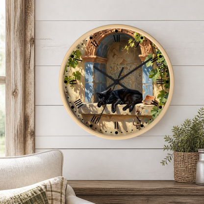 A round wall clock with a light wood frame hangs on a white shiplap wall above a wooden table. The clock face features a scenic illustration of a black cat lying on a windowsill next to an arched window, with green leaves and grapevines, Roman numerals, and black clock hands.
