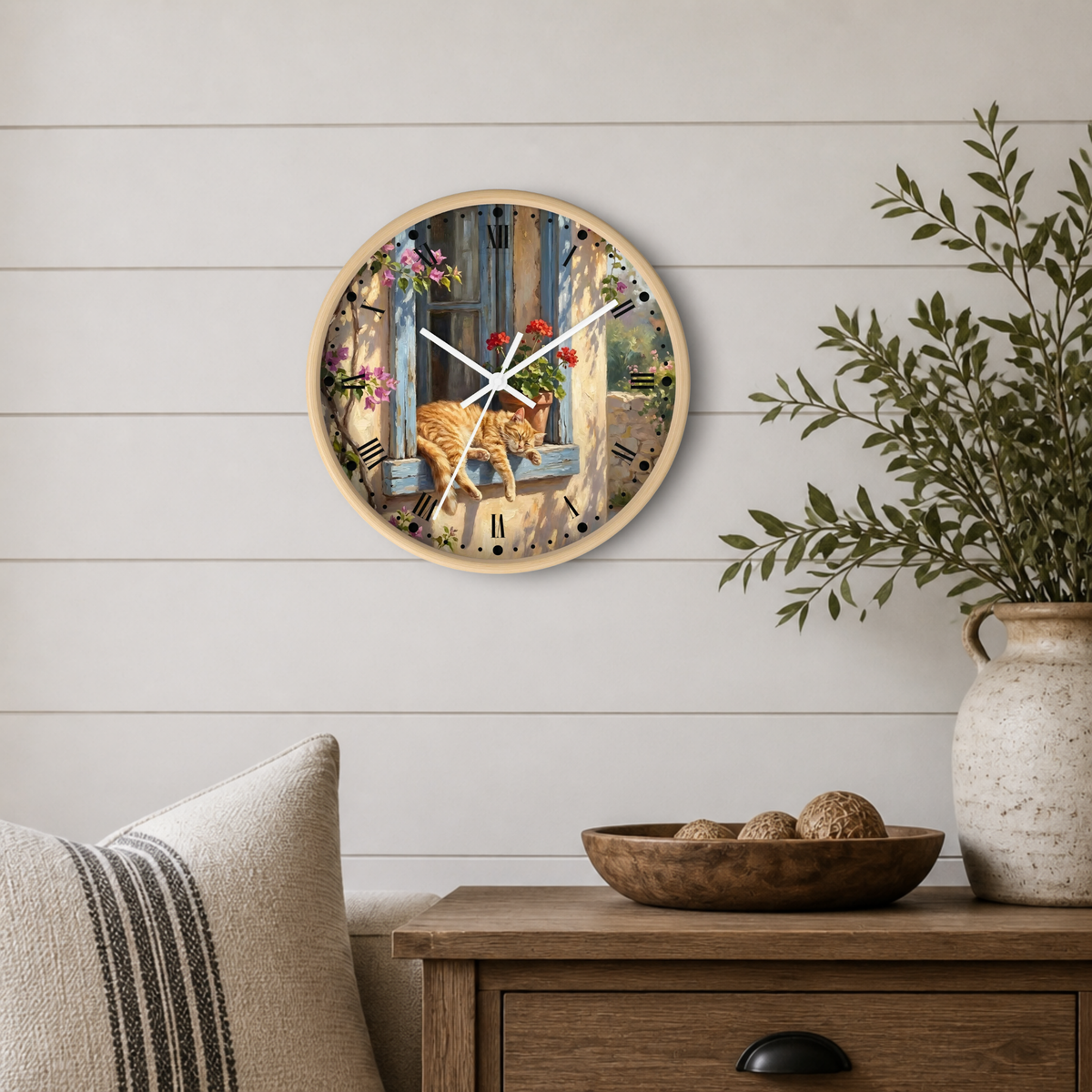 Round wall clock with a rustic illustration of an orange tabby cat sitting on a weathered blue wooden window sill, surrounded by flowers. The clock is mounted on a white shiplap wall above a wooden side table with a plant and decorative bowl.