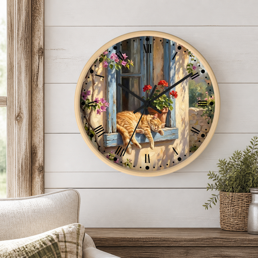 A round wall clock with a light wood frame hangs on a white shiplap wall above a wooden table. The clock face features a detailed illustration of an orange tabby cat sleeping on a blue window sill with red flowers and green leaves.