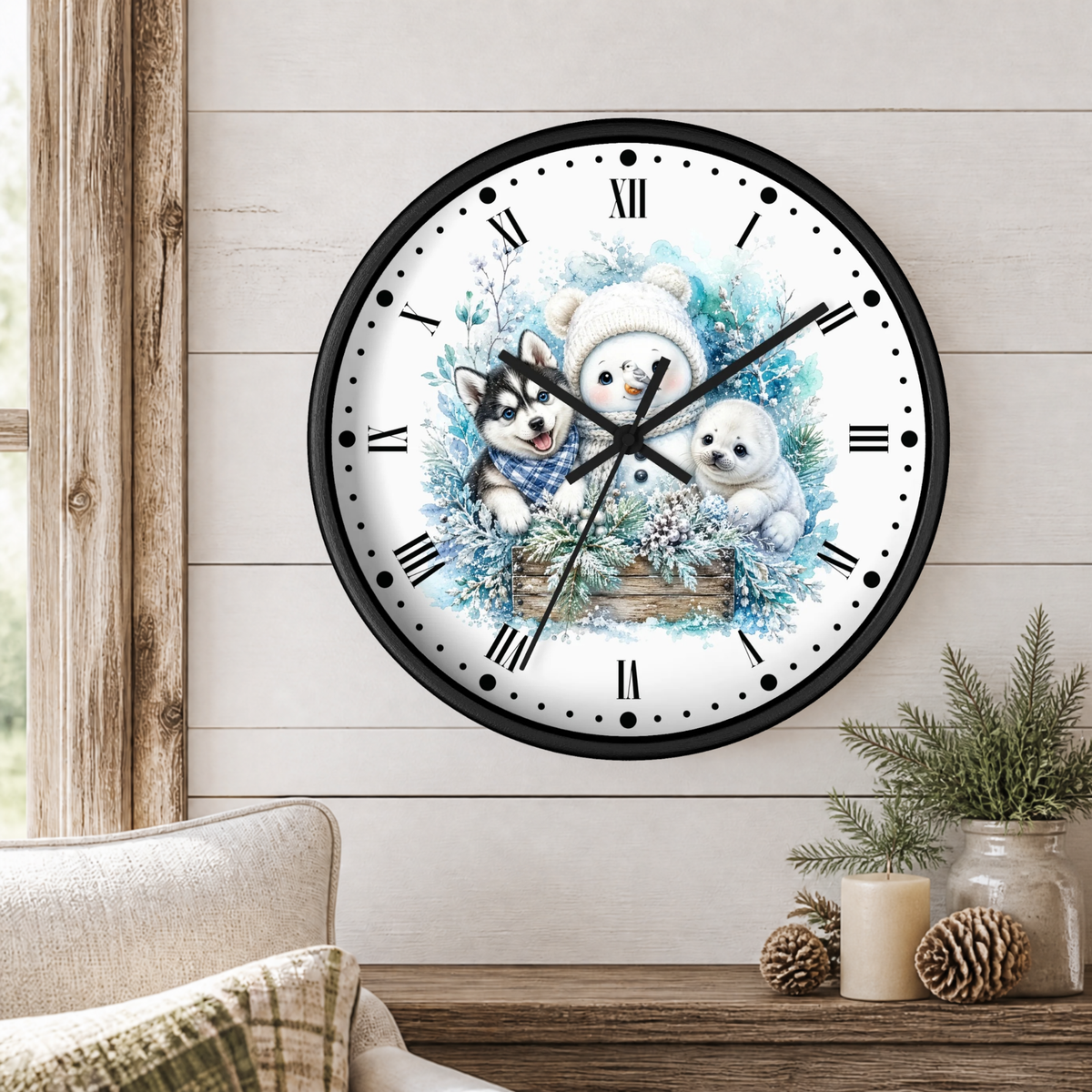 A round wall clock with a black frame and a winter illustration of husky pup, seal and a snowman on the face, mounted on a light wood paneled wall above a wooden table with decorative plants and candles.