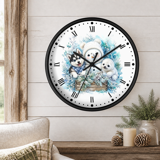 A round wall clock with a black frame and a winter illustration of husky pup, seal and a snowman on the face, mounted on a light wood paneled wall above a wooden table with decorative plants and candles.
