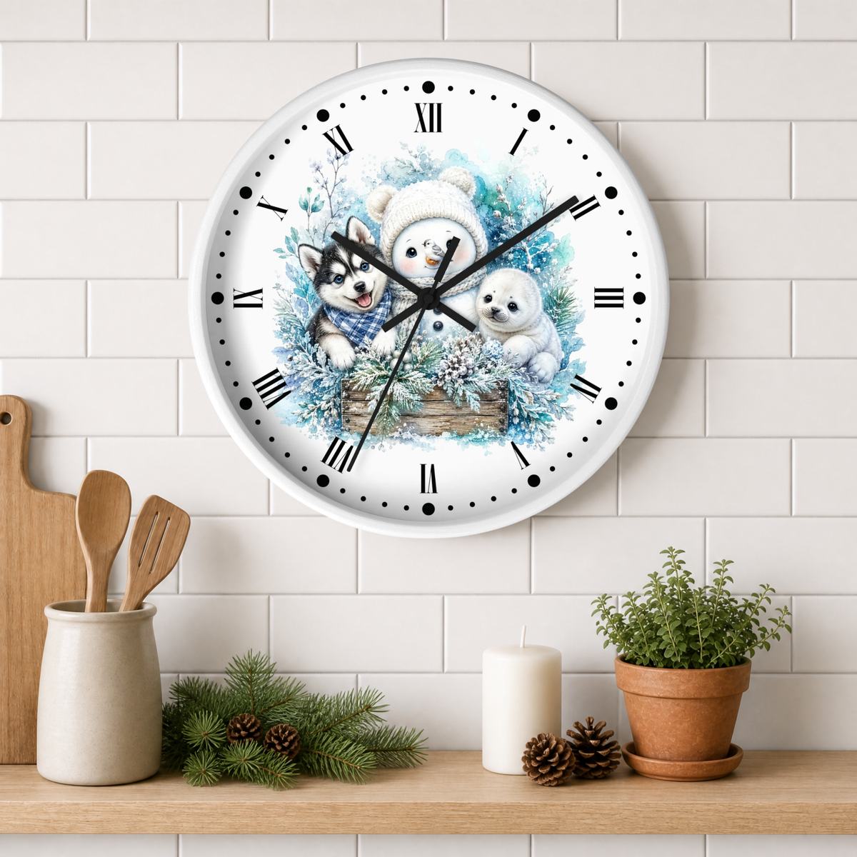 A round wall clock with a white face and black Roman numerals, decorated with an illustration of a husky pup, snowman, and seal in a basket. The clock is mounted on a white subway tile wall above a wooden shelf styled with kitchen utensils, pine branches, a candle, and a potted plant