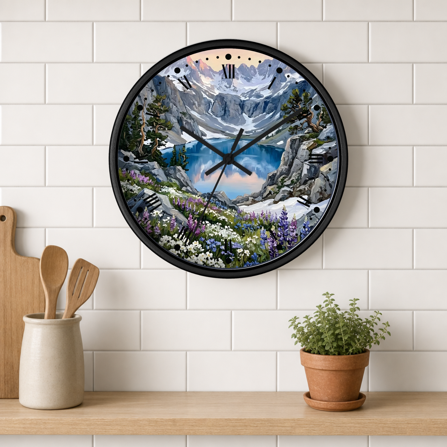 A round wall clock with a black frame and a scenic mountain landscape print, mounted on a white subway tile wall above a wooden shelf with kitchen utensils and a potted plant.