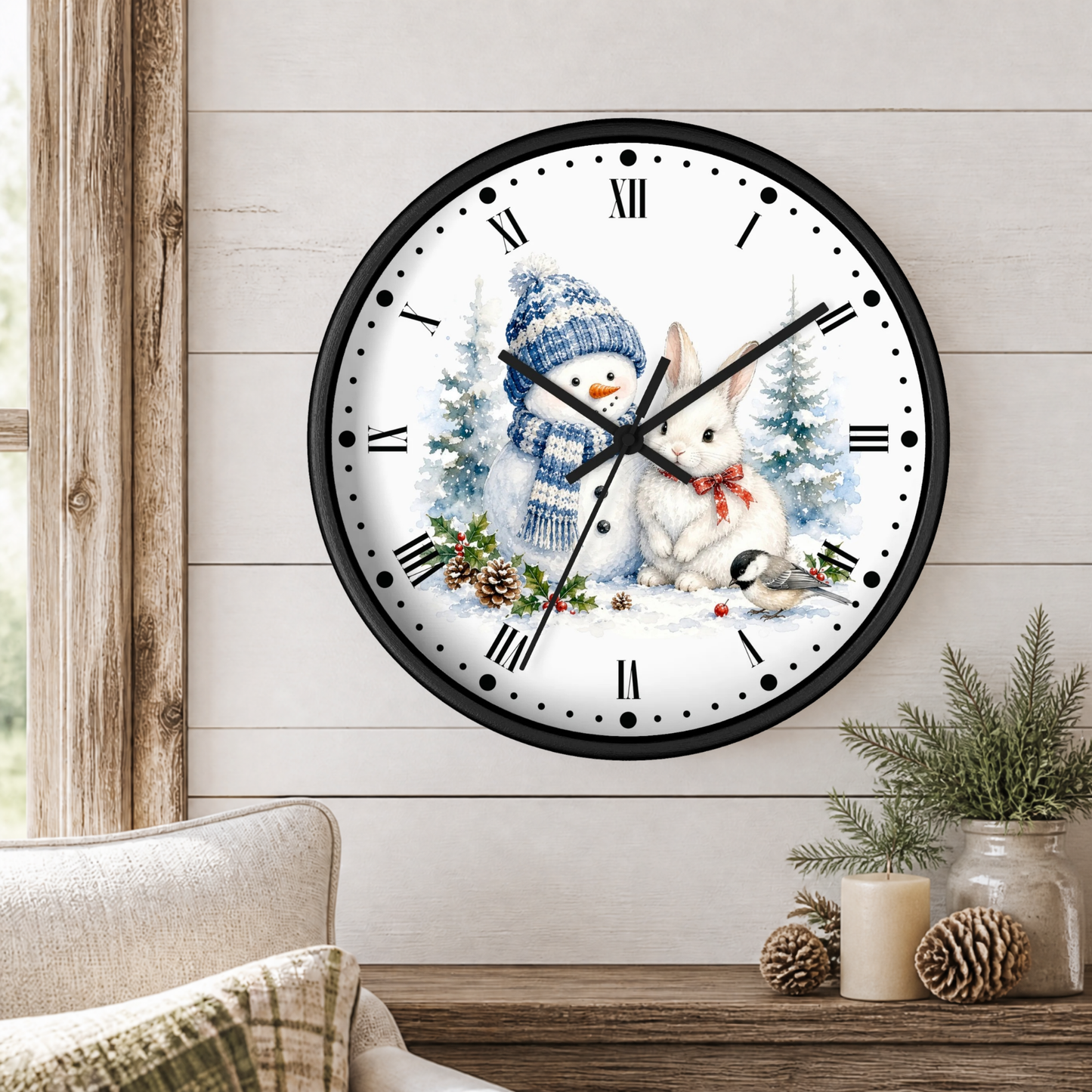 A round wall clock with a black frame and a winter holiday illustration on the face, featuring a snowman in a blue knit hat and scarf and a white rabbit in the snow. The clock is mounted on a light wood paneled wall above a wooden table with pine branches, candles, and pinecones.