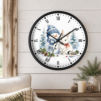 A round wall clock with a black frame and a winter holiday illustration on the face, featuring a snowman in a blue knit hat and scarf and a white rabbit in the snow. The clock is mounted on a light wood paneled wall above a wooden table with pine branches, candles, and pinecones.