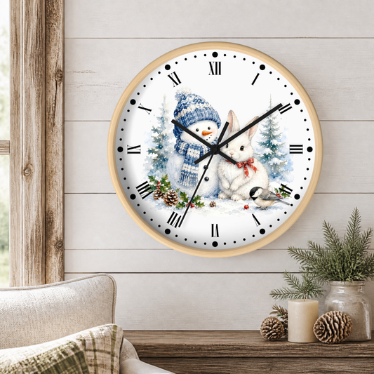 A round wall clock with a light wood frame hangs on a white shiplap wall above a wooden table. The clock face features a winter illustration of a snowman in a blue knit hat and scarf next to a white rabbit with a red bow tie, surrounded by small evergreen trees and pinecones.