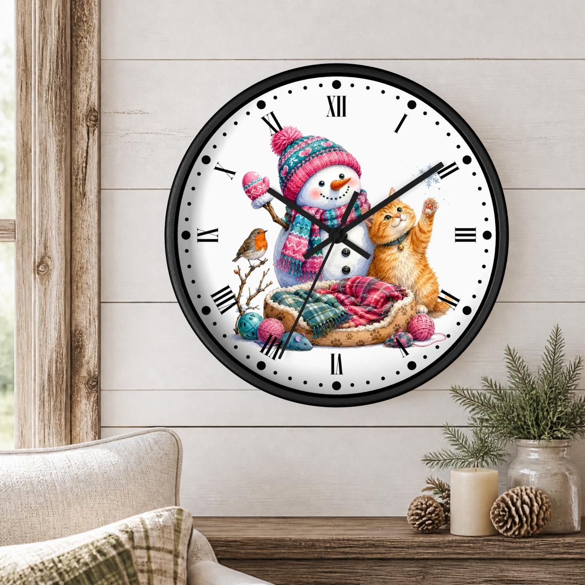 A round wall clock with a black frame and a white face, decorated with a festive winter illustration featuring a snowman in a pink knit hat, a ginger cat, a robin, and colorful yarn balls. The clock is mounted on a light wood-paneled wall above a wooden table with pinecones and a candle.