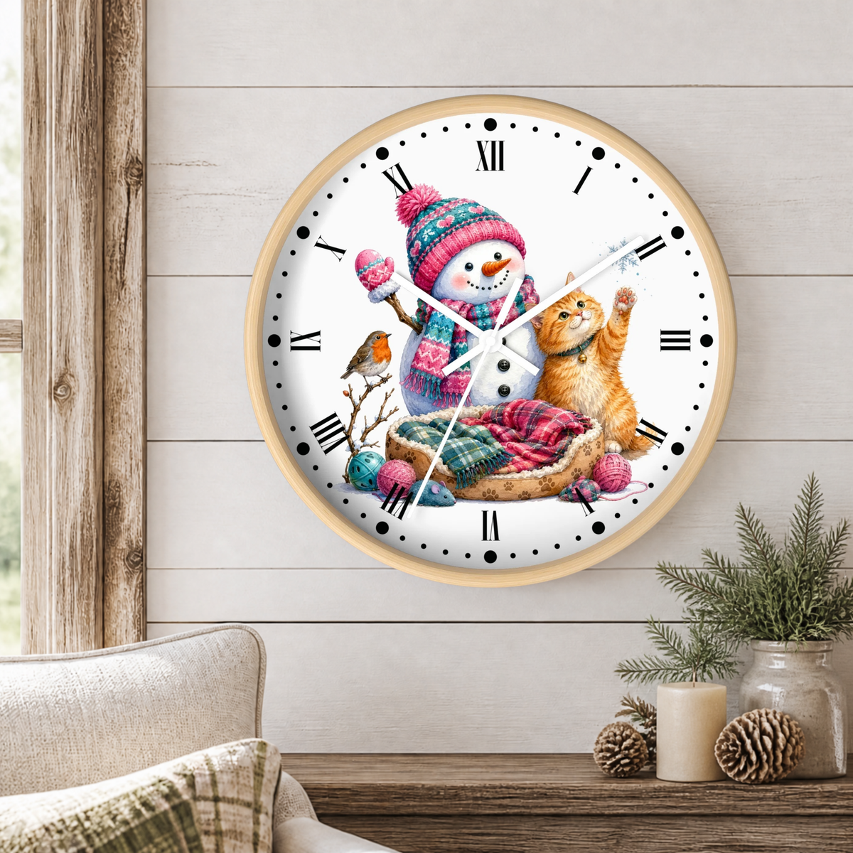 A round wall clock with a light wood frame hangs on a white shiplap wall above a wooden table. The clock face features a whimsical illustration of a snowman wearing a pink and blue knit hat and scarf, a ginger cat, a small bird, and colorful knitted balls.