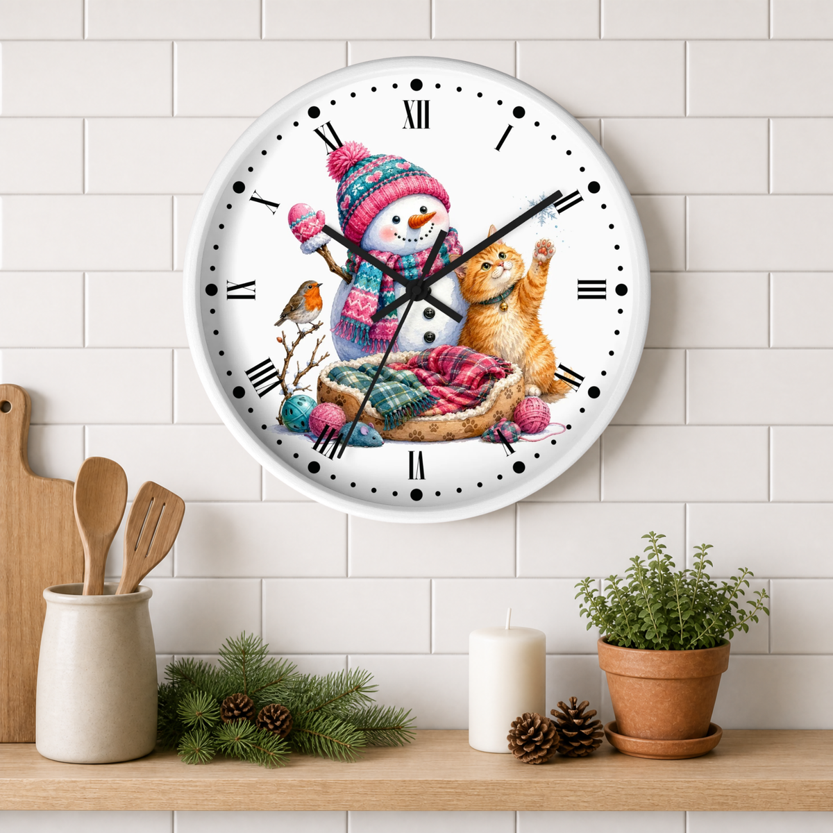 A round wall clock with a white face and black Roman numerals, featuring a colorful illustration of a snowman in a knit hat and scarf, a ginger cat, and a small bird. The clock is mounted on a white subway tile wall above a wooden shelf styled with kitchen utensils, pine branches, a candle, and a potted plant.
