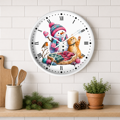 A round wall clock with a white face and black Roman numerals, featuring a colorful illustration of a snowman in a knit hat next to a ginger cat. The clock is mounted on a white subway tile wall above a wooden shelf styled with kitchen utensils, pine branches, a candle, and a potted plant.