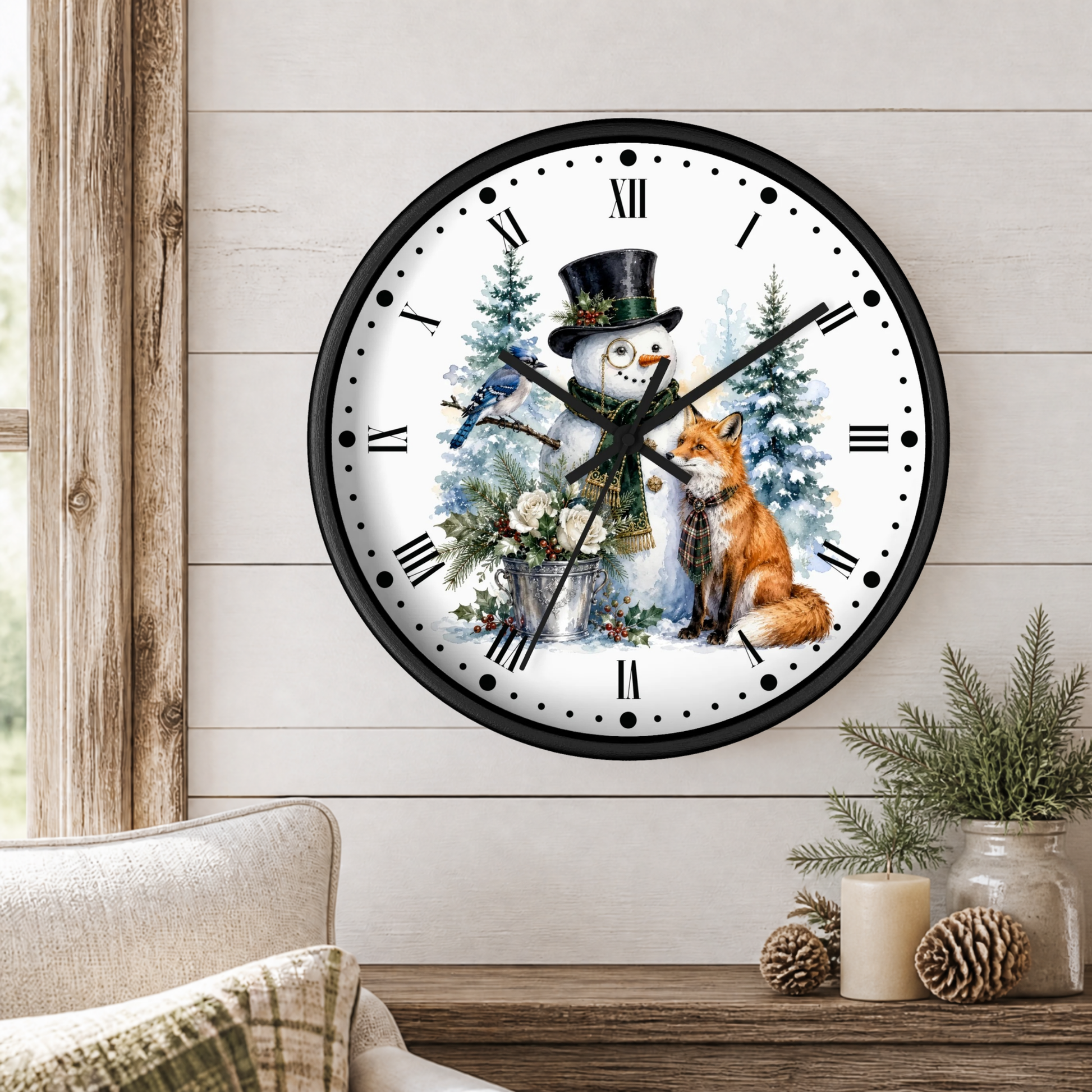 A round wall clock with a winter holiday illustration of a snowman and a fox, mounted on a white wood-paneled wall above a rustic wooden table with candles and pine branches.