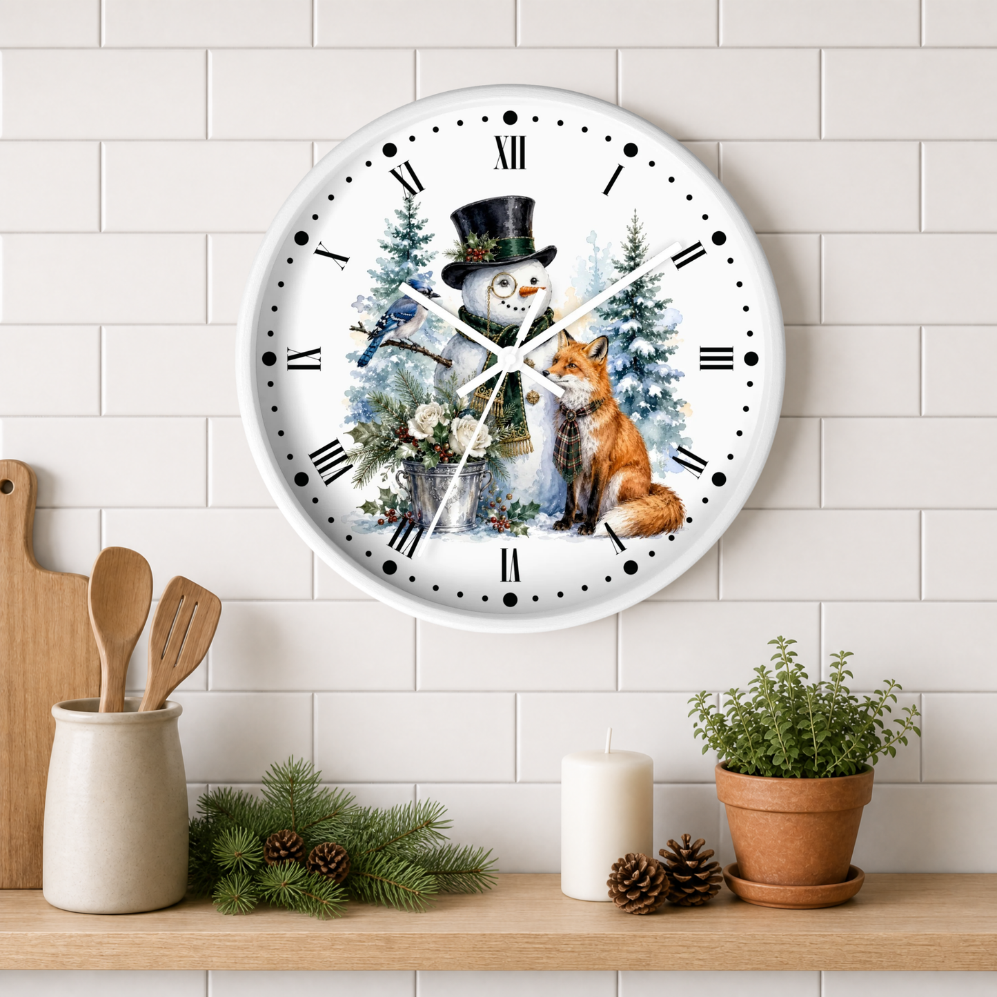 A round white wall clock with a winter holiday illustration featuring a snowman, fox, birds, and evergreen trees. The clock is mounted on a white subway tile wall above a wooden shelf styled with kitchen utensils, pine branches, a candle, and a potted plant.