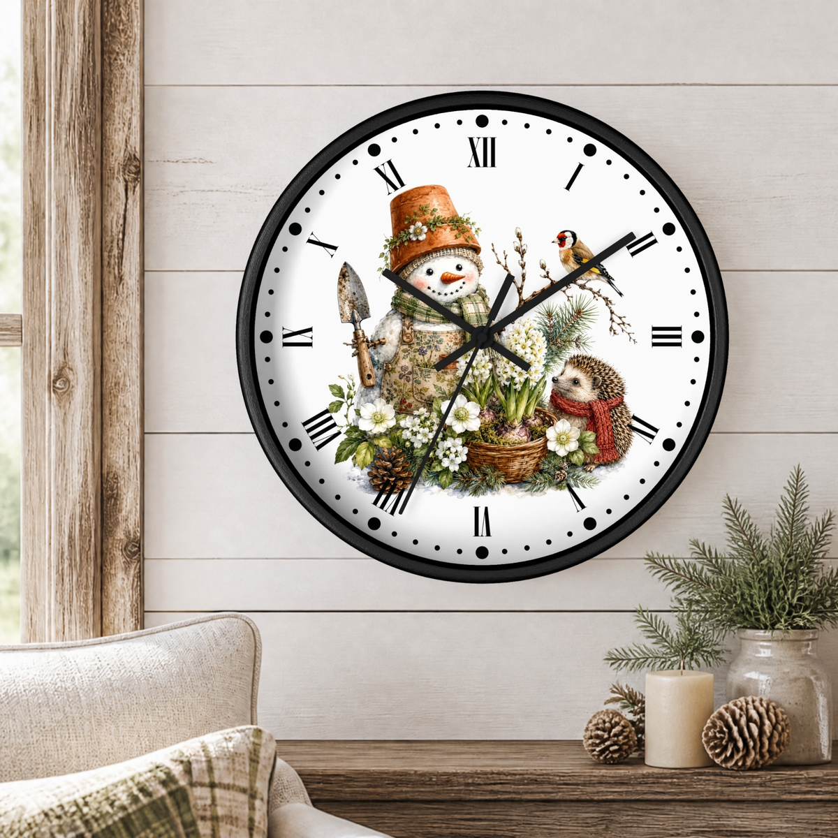 A round wall clock with a black frame and a white face featuring a whimsical winter garden illustration, mounted on a light wood paneled wall above a rustic table with candles and pine branches.