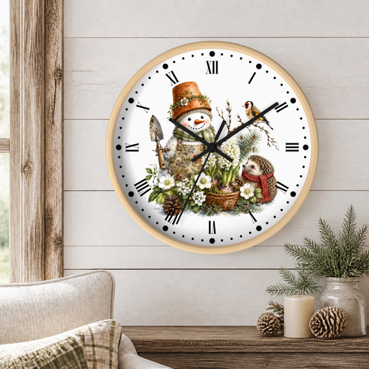 A round wall clock with a light wood frame and a whimsical winter garden illustration with a snowman wearing a plant pot, woodpecker, and hedgehog on the face, mounted on a white shiplap wall above a rustic wooden table with candles and pinecones.