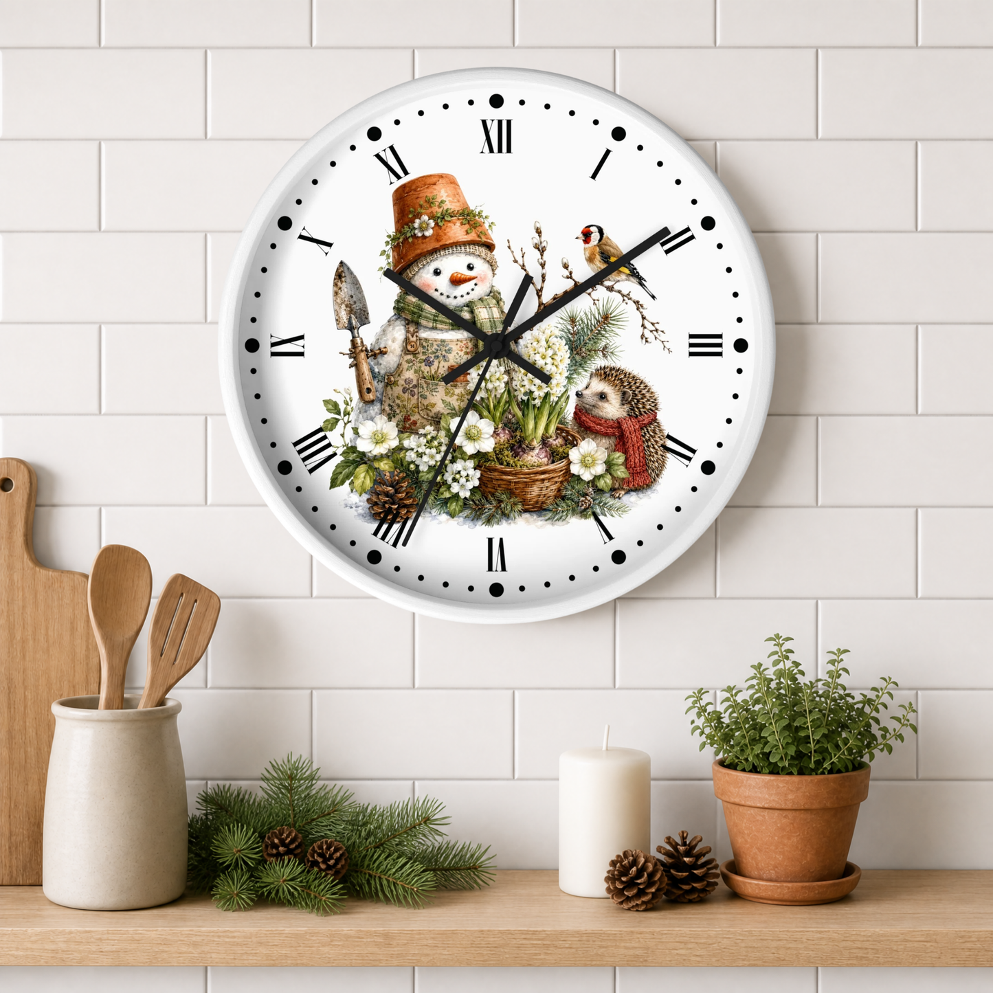 A round wall clock with a white face and black Roman numerals, decorated with a winter garden illustration featuring a snowman, flowers, a bird, and a hedgehog. The clock is mounted on a white subway tile wall above a wooden shelf styled with kitchen utensils, pine branches, a candle, and a potted plant.