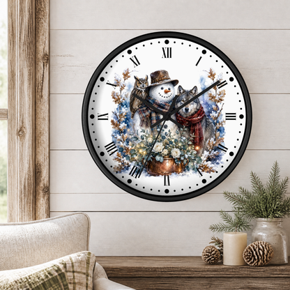 A round wall clock with a black frame and a winter illustration of a snowman, a wolf, and a owls among flowers, mounted on a light wood paneled wall above a table with candles and greenery.