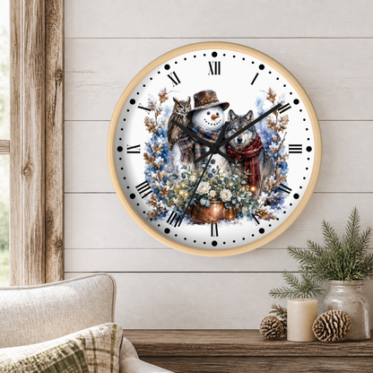 A round wall clock with a light wood frame hangs on a white shiplap wall above a rustic wooden table. The clock face features a detailed illustration of a winter scene with a snowman in a brown hat and red scarf, a gray wolf, owl, and a flower pot with surrounding blue flowers.
