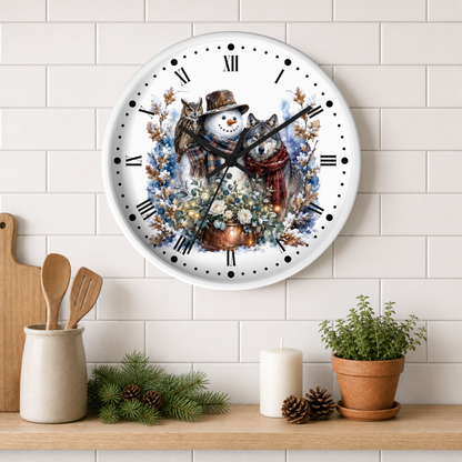 A round wall clock with a white face and black Roman numerals, featuring a winter illustration of a snowman, owl, and a wolf in a cozy winter scene. The clock is mounted on a white subway tile wall above a wooden shelf styled with kitchen utensils, pine branches, a candle, and a potted plant.