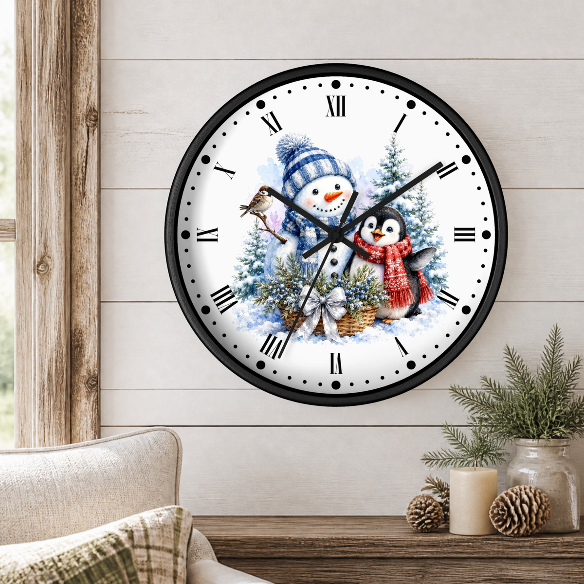 A round wall clock with a black frame and a festive winter illustration of a snowman and penguin under a pine tree, mounted on a light wood paneled wall above a wooden table with candles and greenery.