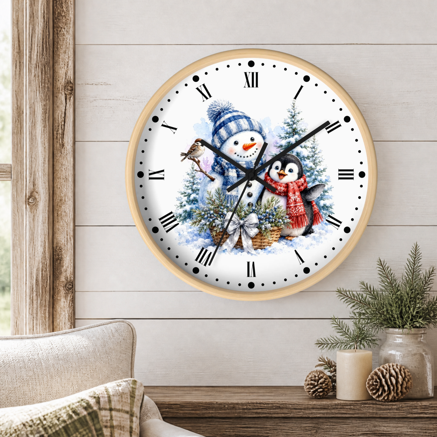 A round wall clock with a light wood frame hangs on a white shiplap wall above a wooden table. The clock face features a winter illustration with a snowman in a blue hat, a penguin in a red scarf, a bird, and evergreen trees.