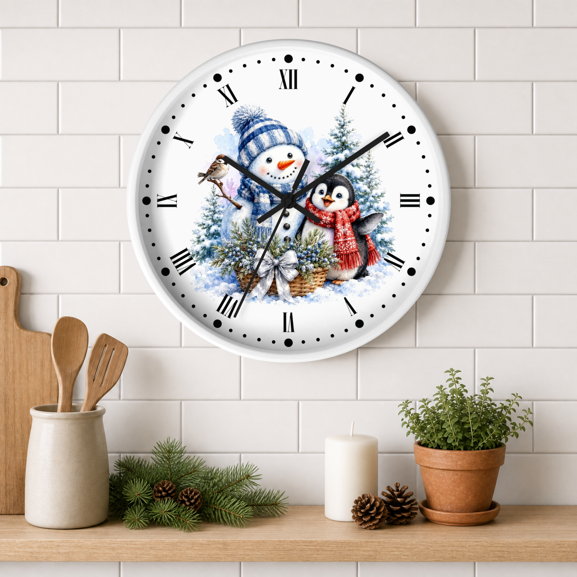 A round wall clock with a white face and black Roman numerals, decorated with a winter holiday illustration of a snowman and a penguin by Christmas trees. The clock is mounted on a white subway tile wall above a wooden shelf styled with kitchen utensils, pine branches, a candle, and a potted plant.