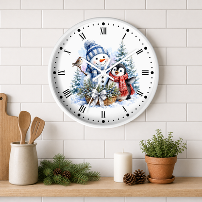 A round wall clock with a winter holiday illustration of a snowman and penguin, mounted on a white subway tile wall above a wooden shelf with kitchen decor and a potted plant.