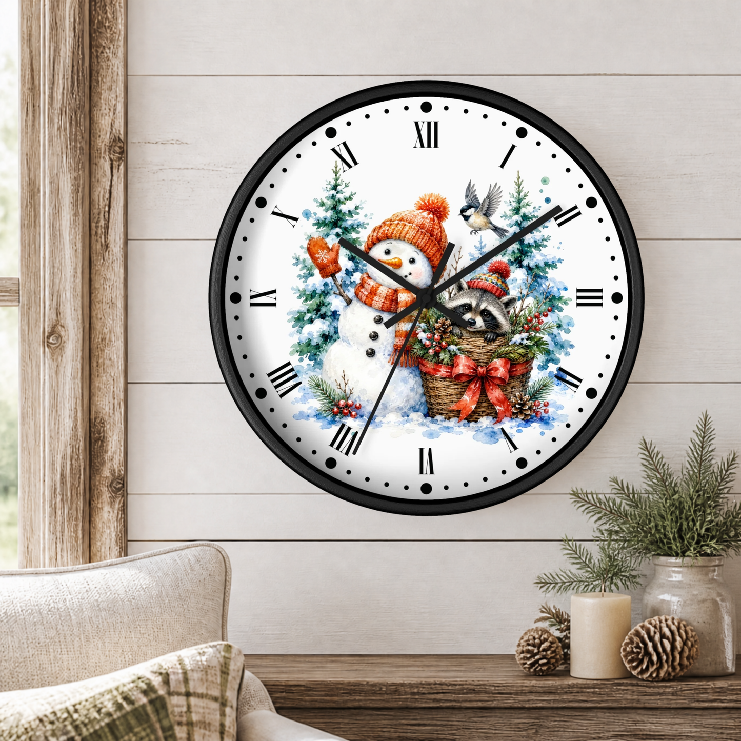 A round wall clock with a black frame and a festive winter illustration featuring a snowman, bird, raccoon, evergreen trees, and a basket of ornaments, shown mounted on a light wood paneled wall above a table with candles and pinecones.
