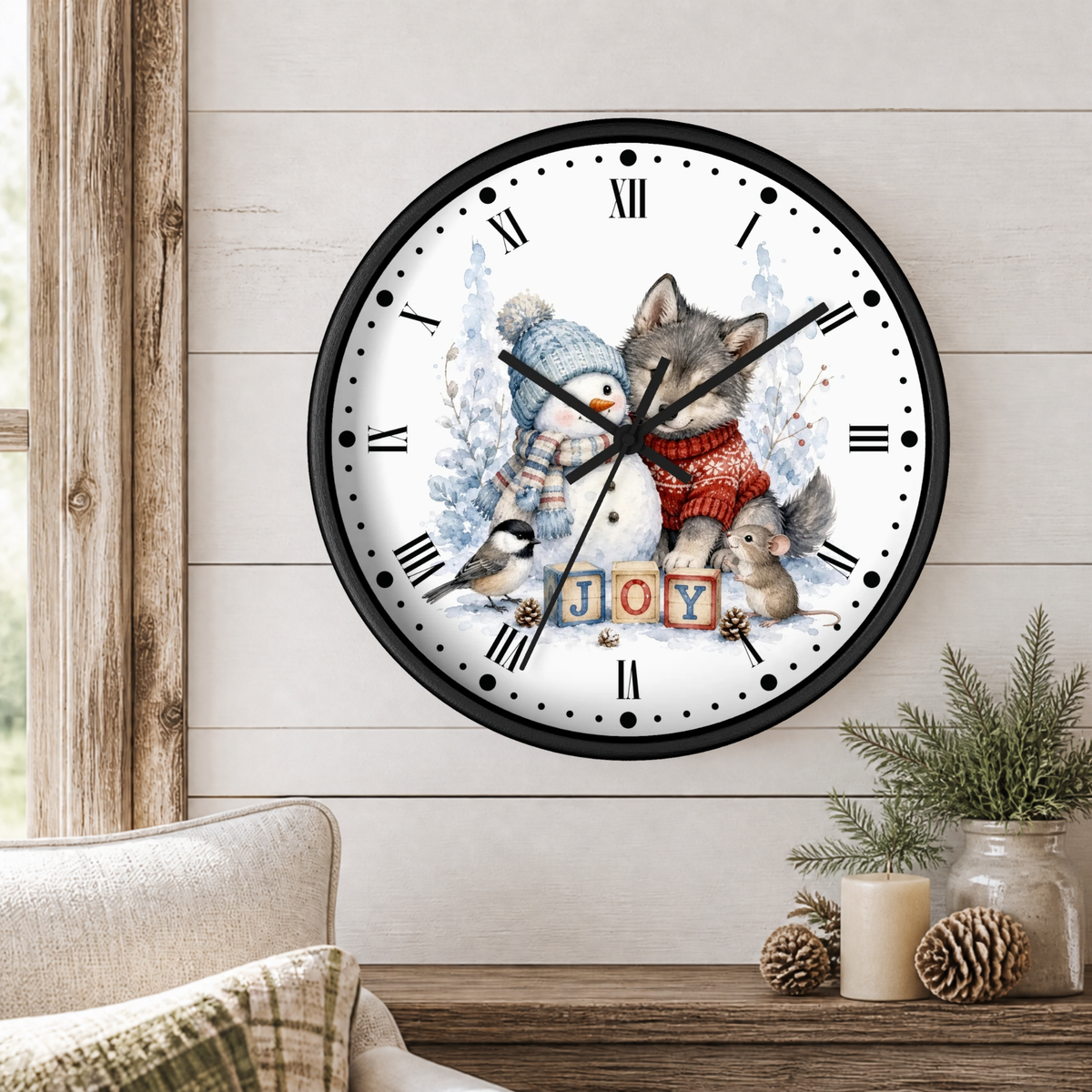 A round wall clock with a black frame and a whimsical winter illustration of a wolf, snowman, bird, and squirrel under the word "JOY," shown hanging on a light wood-paneled wall above a table with candles and greenery.