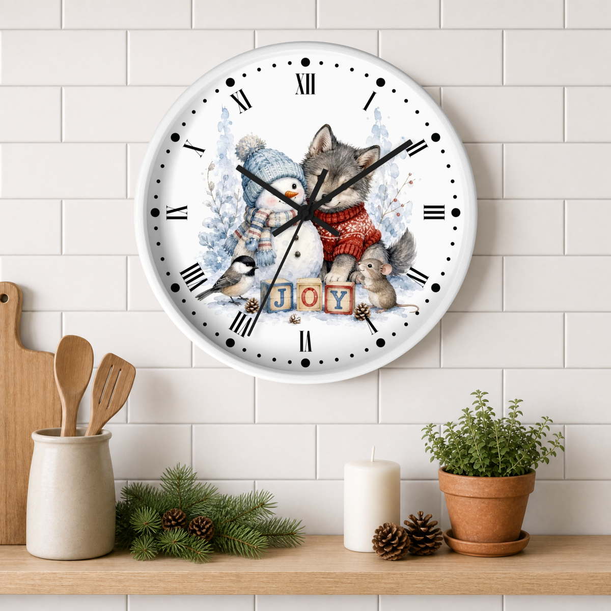 A round wall clock with a white face and black Roman numerals, decorated with a winter illustration of a penguin, a wolf, a squirrel, and the word "JOY" in wooden blocks. The clock is mounted on a white subway tile wall above a wooden shelf styled with kitchen utensils, candles, pinecones, and a potted plant.